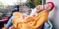 A person sits on a patio chair wrapped in a cozy fleece blanket, while snow falls. Their dog licks their face.