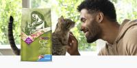 A man smiles as he pets a grey tabby cat in a sunny room; a bag of Purina Cat Chow Naturals Original sits to the right